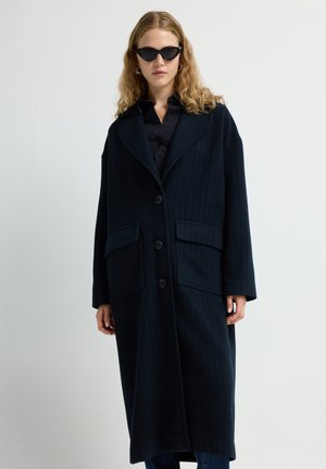 Langer navyblauer Mantel mit vertikalen Streifen, großen Fronttaschen, Knopfverschluss und breiten Revers. Das Modell trägt schwarze Sonnenbrillen und ein dunkles Shirt.