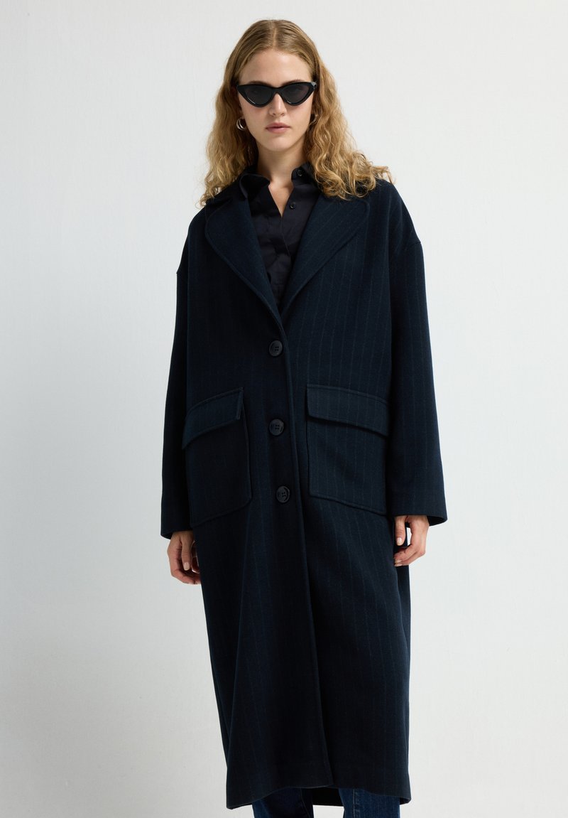 Langer navyblauer Mantel mit vertikalen Streifen, großen Fronttaschen, Knopfverschluss und breiten Revers. Das Modell trägt schwarze Sonnenbrillen und ein dunkles Shirt.