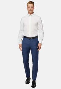 Chemise blanche à col boutonné, associée à un pantalon bleu sur mesure et des chaussures noires à lacets. Design simple, sans motifs ni accents.