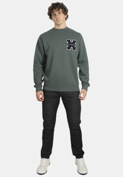 Felpa grigia con collo a giro e tasca frontale, caratterizzata da un design grafico nero. Abbinata a jeans scuri e sneakers bianche.