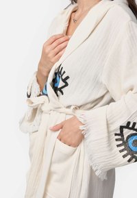 Biela, treskovaná kúpeľňová capa s očným motívom v modrej a čiernej farbe. Obsahuje opasok a bočnú kapsu. Mäkká, textúrovaná látka.