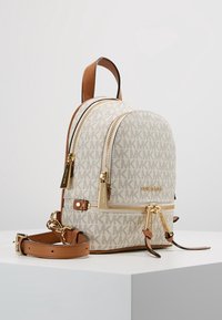 Zaino in tessuto beige con zip dorate, dettagli in pelle marrone e motivo con logo. Presenta una tracolla regolabile e una tasca frontale con zip.