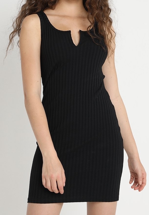 Femme portant une robe noire sans manches côtelée avec une petite encoche en forme de V au niveau de l'encolure, debout devant un fond uni.