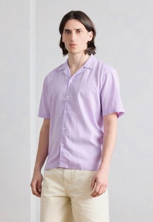 Νέος άνδρας με μεσαίου μήκους σκούρα μαλλιά που φοράει ανοιχτό μωβ κοντομάνικο πουκάμισο με κουμπιά και παντελόνια εκρού, στέκεται σε ουδέτερο φόντο.