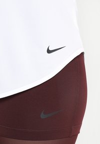 Top atletico bianco con tessuto a rete, orlo curvo e logo Nike nero. Leggings bordeaux con una texture liscia e dettaglio del logo Nike nero.