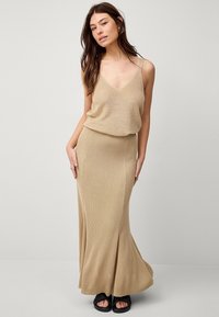 Beige ribbet strik maxi kjole med V-udskæring, tynde stropper og en flydende silhuet, parret med sorte åbne sandaler.