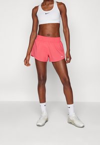 Bermudas rosa de Nike con cinturilla elástica, combinadas con un sujetador deportivo blanco de Nike. La modelo lleva calcetines blancos y zapatillas grises claras.