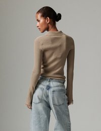 Γυναίκα με μαλλιά σε κότσο, που φοράει εφαρμοστή μπεζ μπλούζα με μακριά μανίκια και ξεφτισμένες μανσέτες, καθώς και ψηλόμεσο ανοιχτό μπλε τζιν, γυρισμένη πλαγίως.