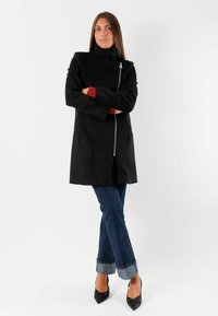 Cappotto nero con colletto alto, chiusura lampo frontale e dettagli rossi sui polsini. Abbinato a jeans in denim e décolleté nere a punta. Texture liscia.