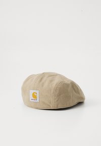 Gorra de color beige hecha de tejido con textura suave. Presenta un parche blanco con un logo amarillo. Tiene una forma redondeada y detalles cosidos.