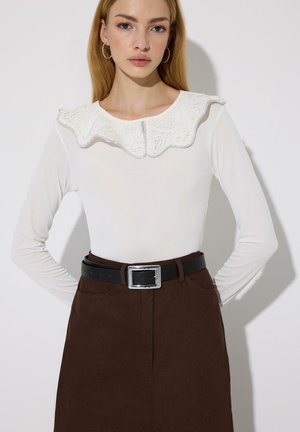 Jeune femme portant un haut blanc à manches longues avec un large col en dentelle, une jupe marron taille haute et une ceinture noire avec boucle en argent.