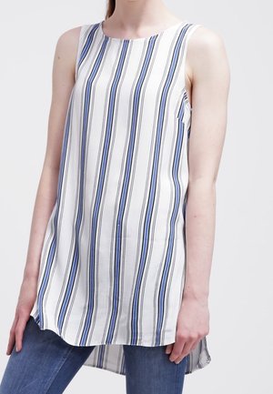 Woman wearing sleeveless white tunic with vertical blue and black stripes, paired with blue jeans, against a plain background.