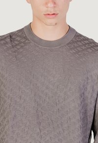 Pull en tricot gris avec un motif texturé de lettres, col rond à côtes et légère brillance, mettant en valeur les détails du tissu.