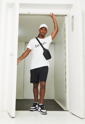 Svart shorts, hvit T-skjorte med "LOS" tekst, svart crossbody-veske, og svart/hvite sneakers. Står i en døråpning med en minimalistisk bakgrunn.