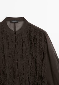Blusa marrone scuro con ruches verticali testurizzate, colletto rotondo, bottoni frontali e maniche lunghe trasparenti.