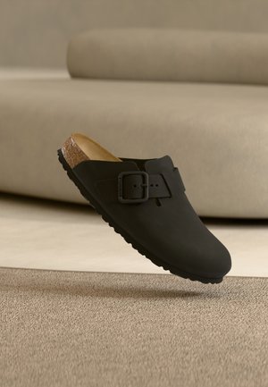 Sort slip-on klodsko med justerbar spænde og korksål, vist svævende over beige tekstureret tæppe og polstret sofa i baggrunden.