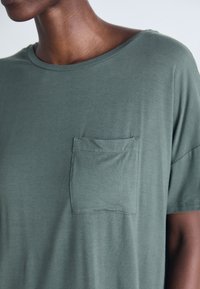 Närbild av person som bär en dämpad grön kortärmad skjorta med en vänster bröstficka och rund halsringning.