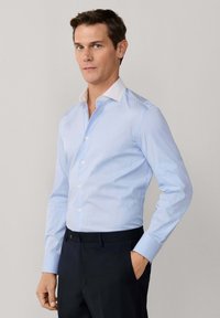 Mann trägt hellblaues Hemd mit weißem Kragen und dunkle Hose, steht mit einer Hand in der Tasche vor neutralem Hintergrund.