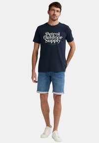 Námořnická modrá bavlněná trička s potiskem "Petrol Outdoor Supply" v krémové barvě. Spárováno s světle modrými džínovými šortkami a bílými teniskami.