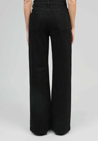 Personne portant un pantalon noir taille haute à jambes larges avec un motif à pois subtil, vu de dos contre un fond gris uni.
