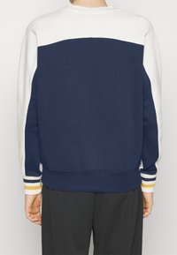 Tmavě modrá mikina s bílou horní částí, s proužkovanými akcenty v šedé, žluté a bílé na manžetách. Měkká tkanina a uvolněný střih.