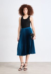 Top aderente nero con paillettes abbinato a una gonna midi plissettata color teal e tacchi neri con cinturini. La modella tiene una piccola pochette nera.