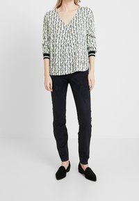 Blouse à manches longues en V sur un fond clair avec un motif de feuilles noires, associée à un pantalon noir slim et des mocassins noirs plats.