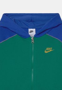 Sudadera con cremallera en verde y azul, con un logo dorado de Nike, que presenta una textura suave, bloques de color en contraste y detalles de costura visibles.