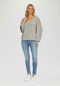 Grijze V-hals sweatshirt met lange, iets opgeblazen mouwen en een relaxte pasvorm, gecombineerd met lichtblauwe skinny jeans en witte sneakers.