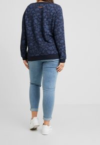 Sudadera azul marino con patrón de hojas, escote redondeado, puños y dobladillo acanalados. Combinada con jeans ajustados azul claro y zapatillas blancas.
