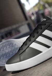 Basket Adidas noire et blanche avec semelle blanche et trois bandes blanches, inclinée pour montrer le côté et la semelle à motifs.