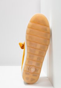 Semelle d’une chaussure jaune avec une semelle extérieure en caoutchouc translucide et un motif de grip texturé, positionnée à la verticale contre un coin blanc.