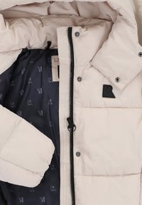 Een pufferjack in een crèmekleur met een ritssluiting aan de voorkant en drukknoopjes. Het interieur heeft een marineblauwe voering met een subtiel logopatroon. Zwarte logopatch op de mouw.