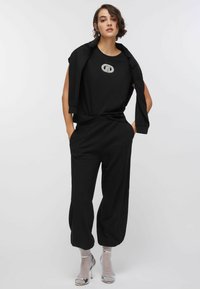 Ung kvinde iført sort tøj med en ærmeløs top, vide bukser, jakke hængende over skuldrene samt sølvfarvede høje hælsandaler med sokker.