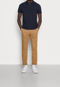 Polo shirt blu navy, chinos beige, sneakers bianchi e calzini bianchi. La polo presenta un colletto e un'apertura a due bottoni; i chinos hanno i risvolti.