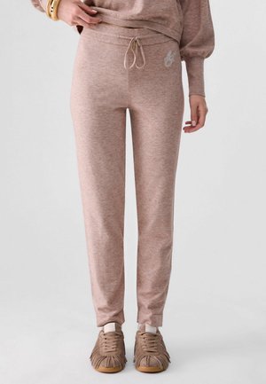 Femme portant un pantalon de survêtement marron clair ajusté avec taille à cordon et des baskets assorties à franges, debout devant un fond uni.
