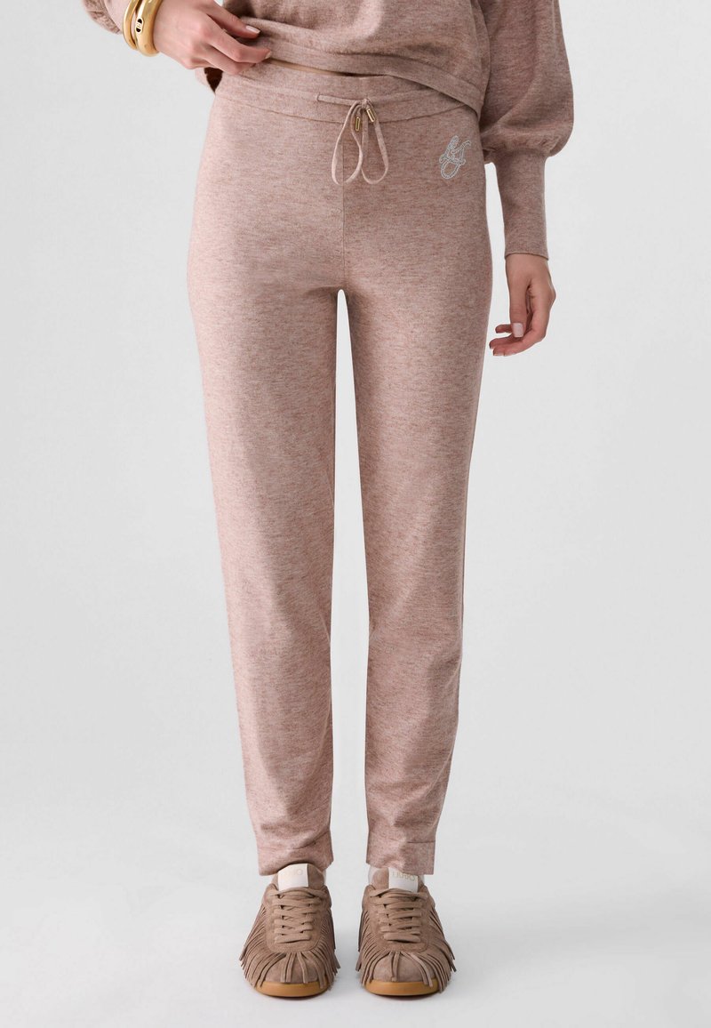 Femme portant un pantalon de survêtement marron clair ajusté avec taille à cordon et des baskets assorties à franges, debout devant un fond uni.
