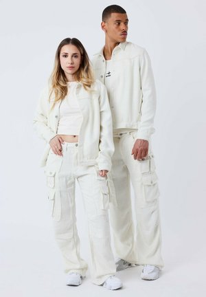 Due persone indossano abiti coordinati di un bianco sporco testurizzato. L'outfit della donna comprende una giacca a maniche lunghe e pantaloni cargo, mentre l'uomo indossa una maglietta a maniche corte e una giacca.