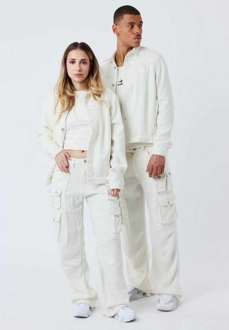 Deux personnes portent des vêtements assortis de couleur écru texturée. La tenue de la femme comprend une veste à manches longues et un pantalon cargo, tandis que l'homme porte un haut à manches courtes et une veste.