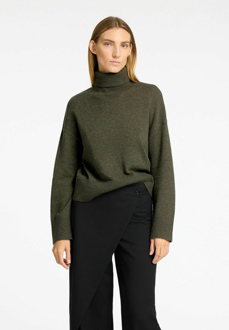 Dunkelgrüner Strickrollkragenpullover mit lockerer Passform, gerippten Bündchen und Saum. Kombiniert mit schwarzen, hochtaillierten, weit geschnittenen Hosen.