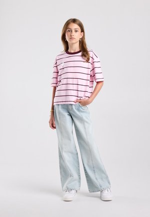 Jonge vrouw staat met één hand in de zak, draagt een roze gestreept T-shirt, lichtblauwe wijde jeans en witte sneakers tegen een lichte achtergrond.