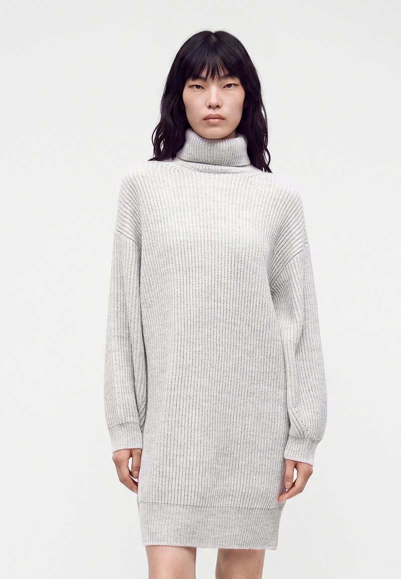 Femme aux cheveux noirs portant une robe pull en maille côtelée gris clair à col roulé, debout devant un fond blanc uni.