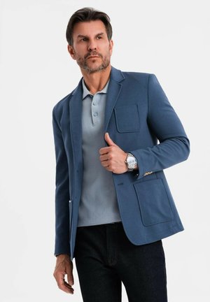 Uomo con barba chiara che indossa un blazer blu, una polo azzurra chiara, pantaloni scuri e un orologio in metallo contro uno sfondo bianco uniformemente.
