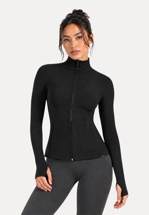 Mujer vestida con una chaqueta negra ajustada con cremallera y agujeros para los pulgares y leggings gris oscuro, de pie frente a un fondo blanco liso.