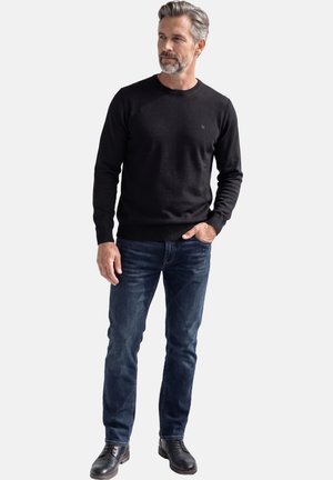 Mann mit grauem Haar und Bart, der einen schwarzen Pullover, dunkelblaue Jeans und schwarze Lederschuhe trägt, steht mit einer Hand in der Tasche.