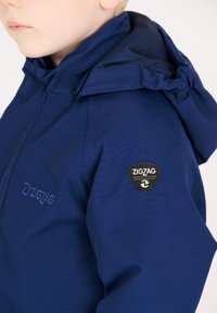 Marineblauwe jas met capuchon, met een textuurstof, geborduurd logo op de borst en een badge op de mouw.
