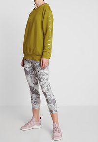 Sweat-shirt vert olive avec le texte "JUST DO IT" sur la manche, associé à des leggings gris à motif de feuilles. Des chaussures de sport roses complètent la tenue.