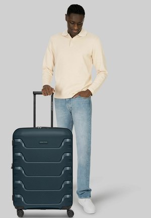 Uomo con camicia beige a maniche lunghe e jeans, in piedi con la mano sulla maniglia di una grande valigia rigida blu scuro con rotelle.