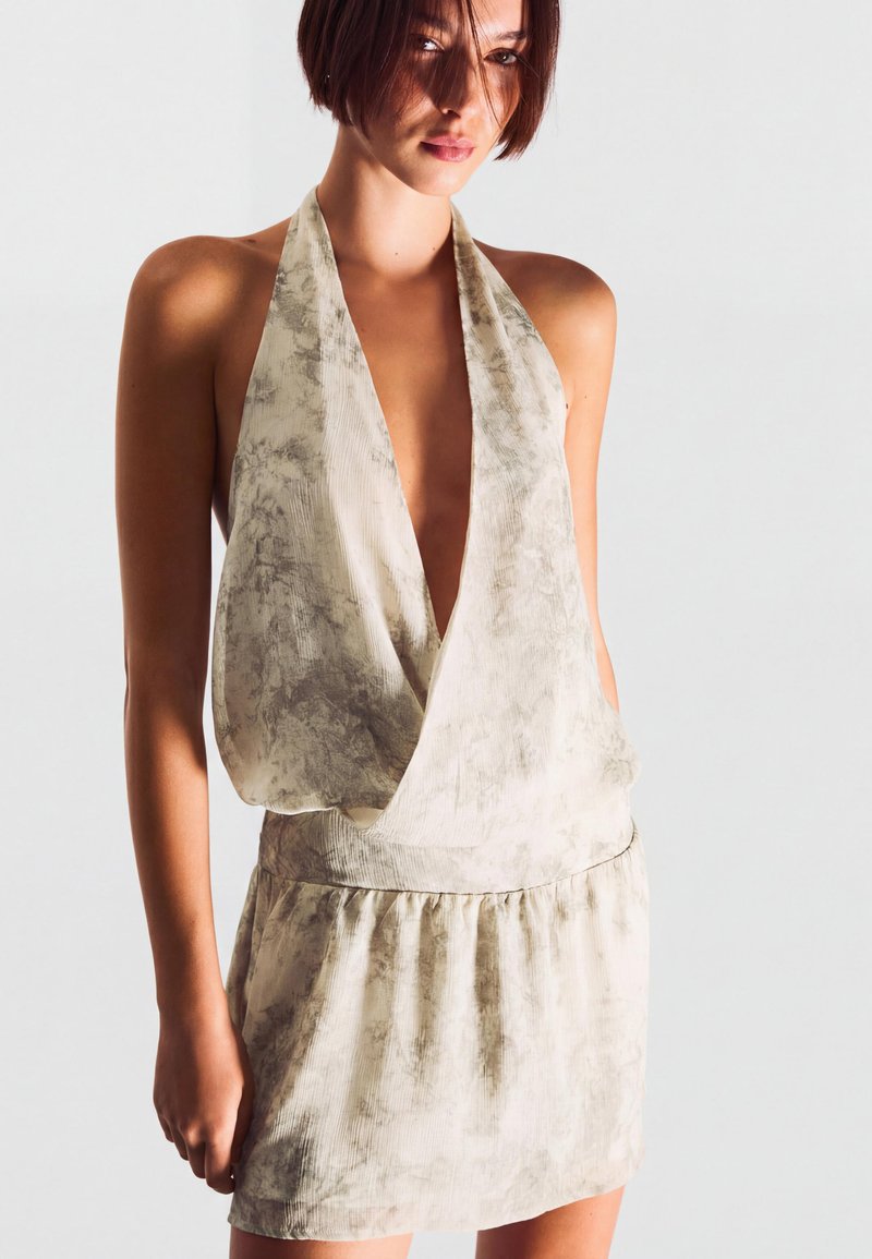 Femme portant une robe sans manches, beige clair texturée, à col licou profond en V, debout devant un fond clair uni.