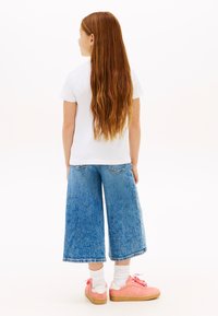 Παιδί με μακριά κόκκινα μαλλιά που φοράει λευκό μπλουζάκι, φαρδιά cropped τζιν παντελόνια, λευκές κάλτσες και ροζ αθλητικά παπούτσια, στέκεται με την πλάτη.